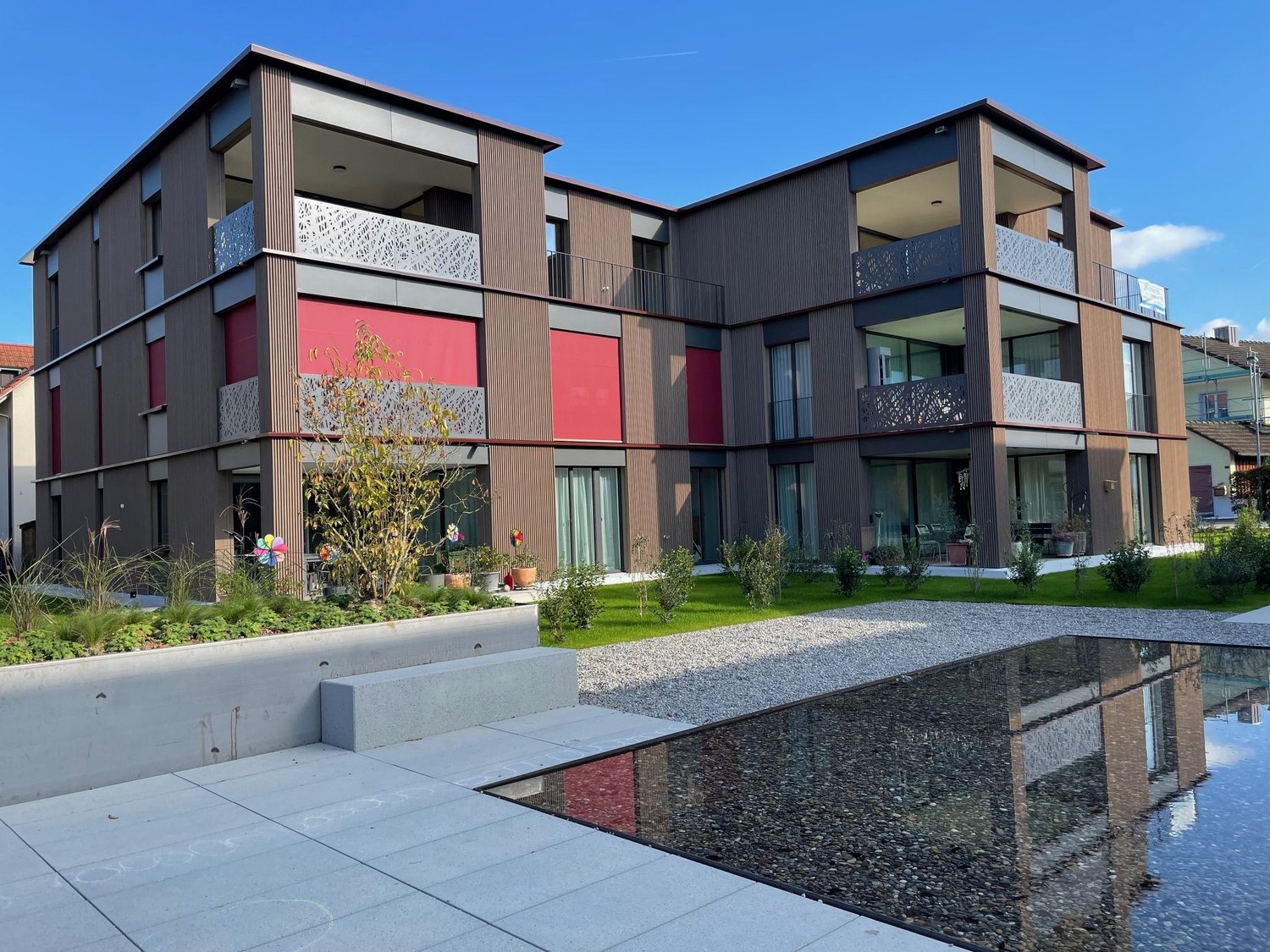 Modern multi-story building, brown exterior, red accents, multiple balconies, landscaped front yard with water feature