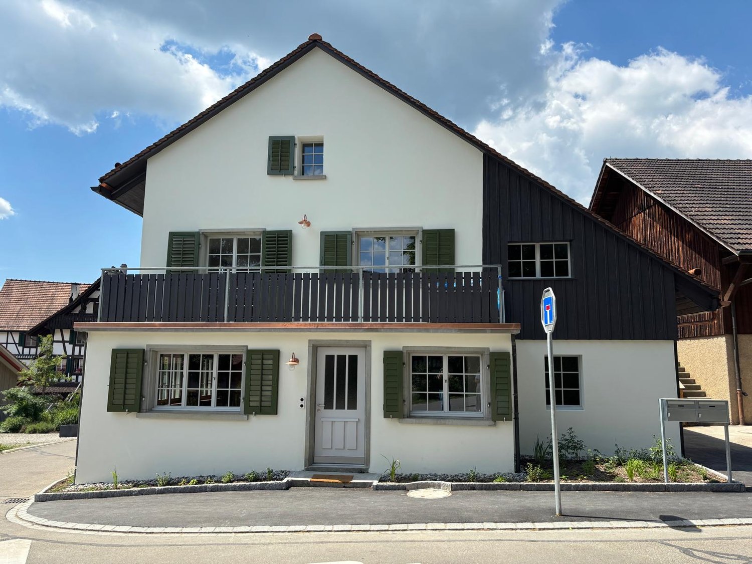 2-story house with white exterior walls, green shutters, and a balcony on the second floor. The house has a tiled roof and is surrounded by a paved driveway or walkway.