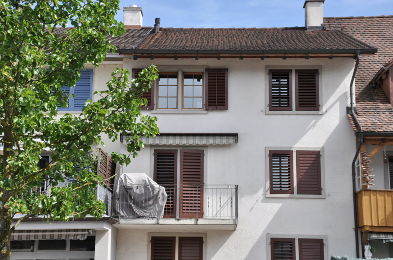 Three-story building, white walls, brown roof, brown shutters, balcony on second floor, awnings