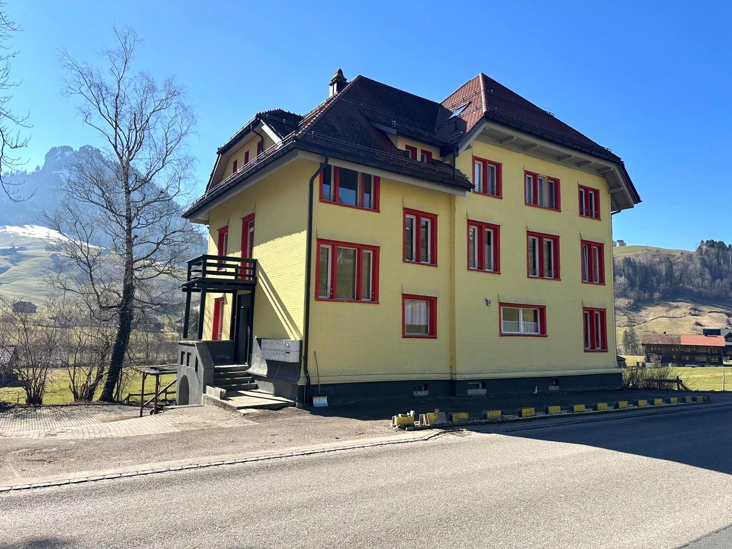 3 story building, yellow exterior, red framed windows, steep roof, rural landscape