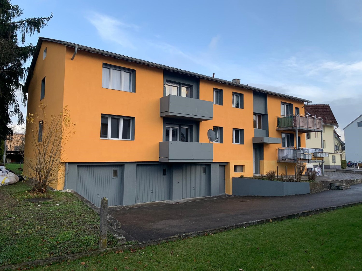 Three-story building, yellow walls, gray roof, windows, balconies, ground floor parking, nearby sidewalk