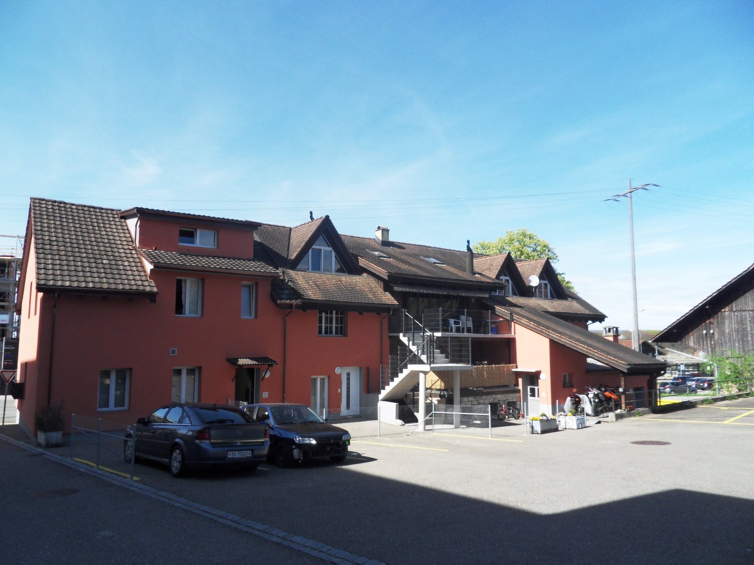 red two-story house with brown roof, two balconies, stairs, plants, two cars parked in front, one motorcycle parked