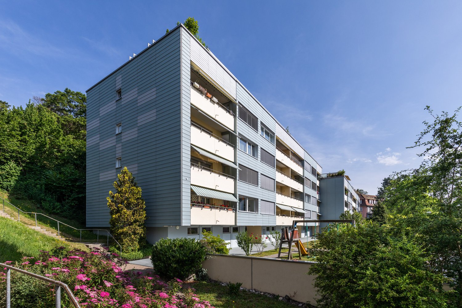 Gray modern apartment building, multiple balconies, fenced playground with slides, lush greenery