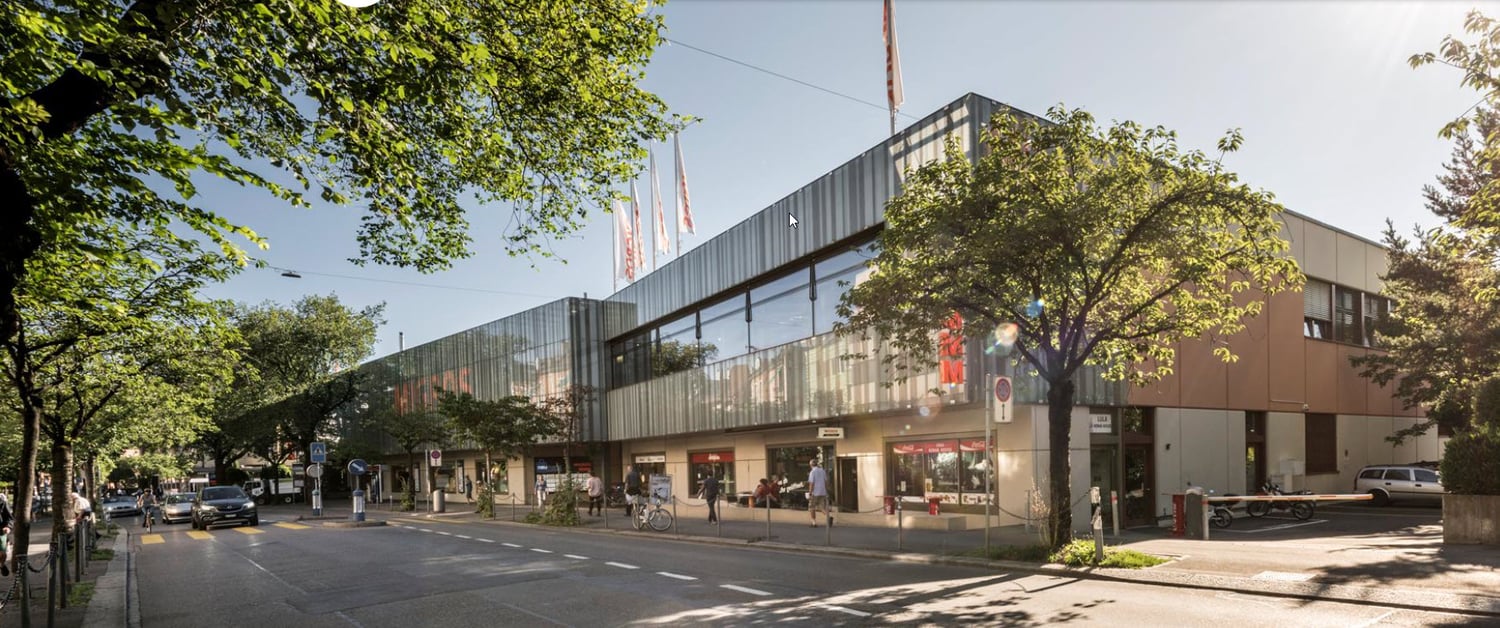 A multi-story commercial building with a glass facade, surrounded by trees and pedestrians on the street. The building appears to house various shops and businesses.