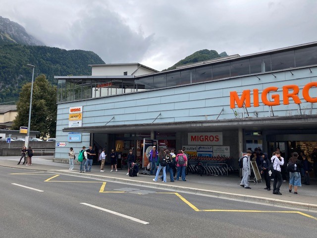 Front entrance of the Migros store with glass walls and doors, many people are entering