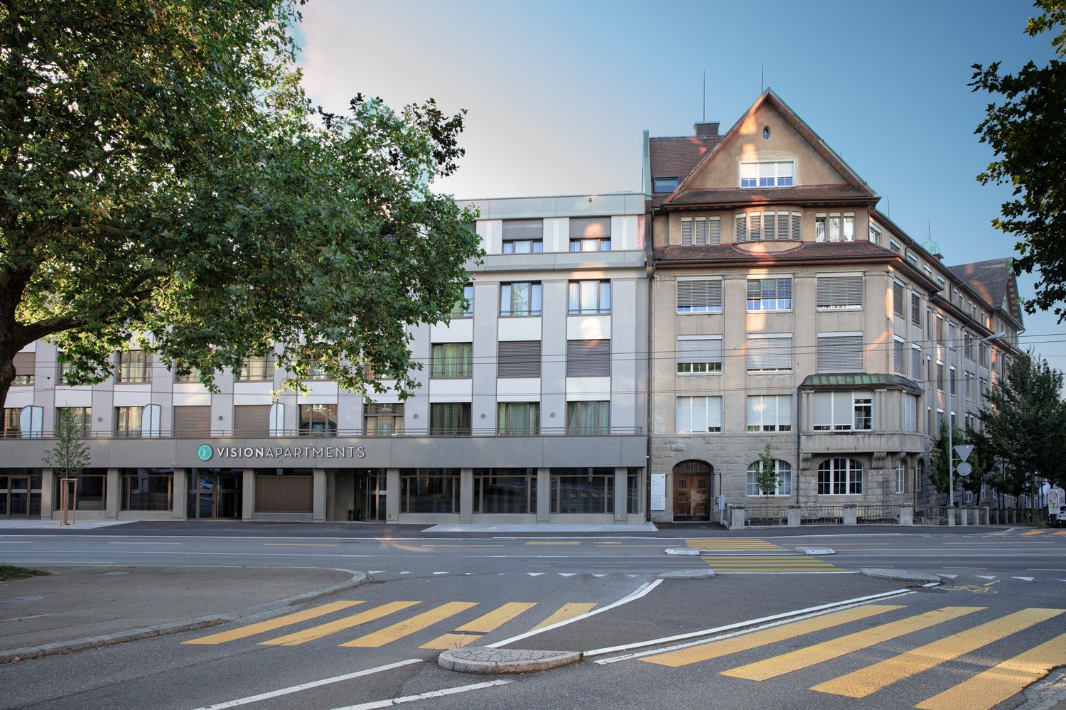 Two-storey building with multiple windows and entrance on street, tree in front, sign reading VISIONAPARTMENTS