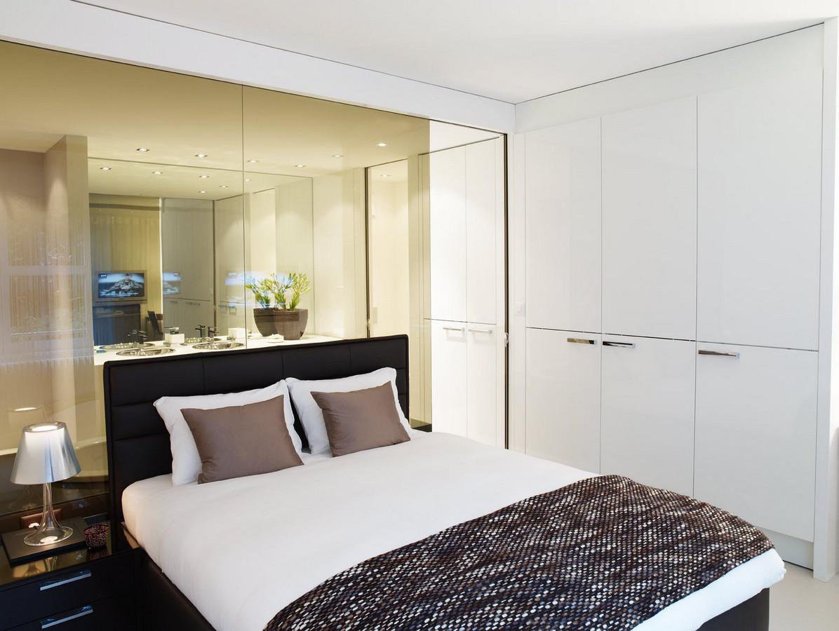 white bedroom, double bed, two pillows, white walls, full mirror on wall, cabinet with drawers, nightstand with lamp