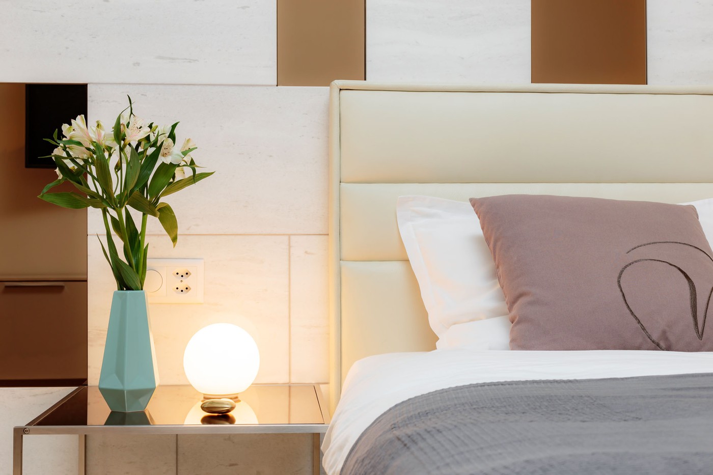 a bed with white bedding, cream-colored pillow and teal pillow, nightstand with lamp and flower vase, white wall, wall socket with plug