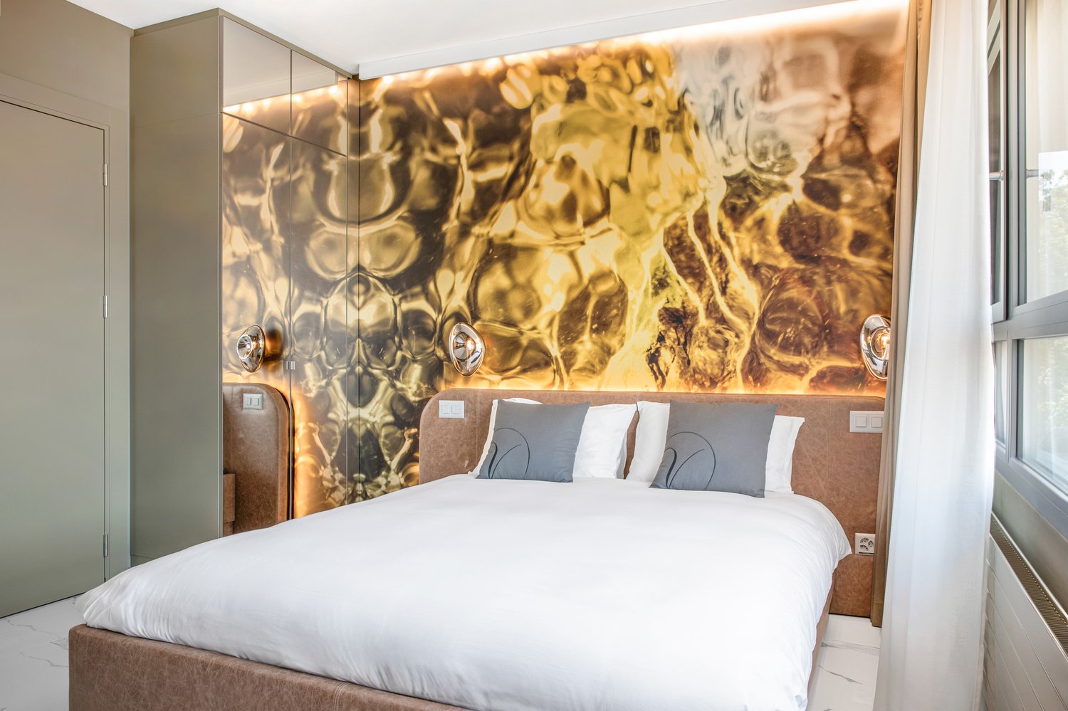 Double bed with white linen, two grey pillows, golden wall mural, large windows