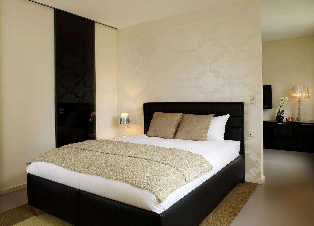 double bed with black headboard, beige blanket, two beige pillows, two white pillows, carpeted floor, two bedside lamps, black wall paneling, patterned wallpaper, adjacent dressing room, small flower vase on counter, TV