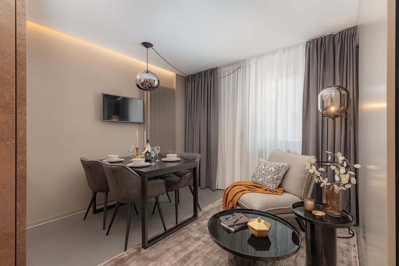 A dining area with a set of four chairs, a coffee table, a TV mounted on the wall, and a small couch with a coffee table.