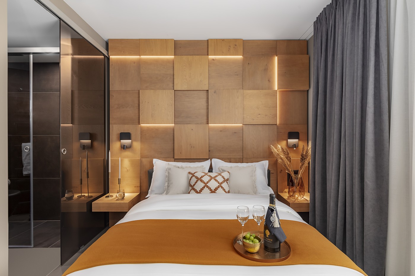 Modern bedroom with a king size bed, two bedside tables, decorative bottles, wine glasses, two nightstand lamps, wooden walls and shower