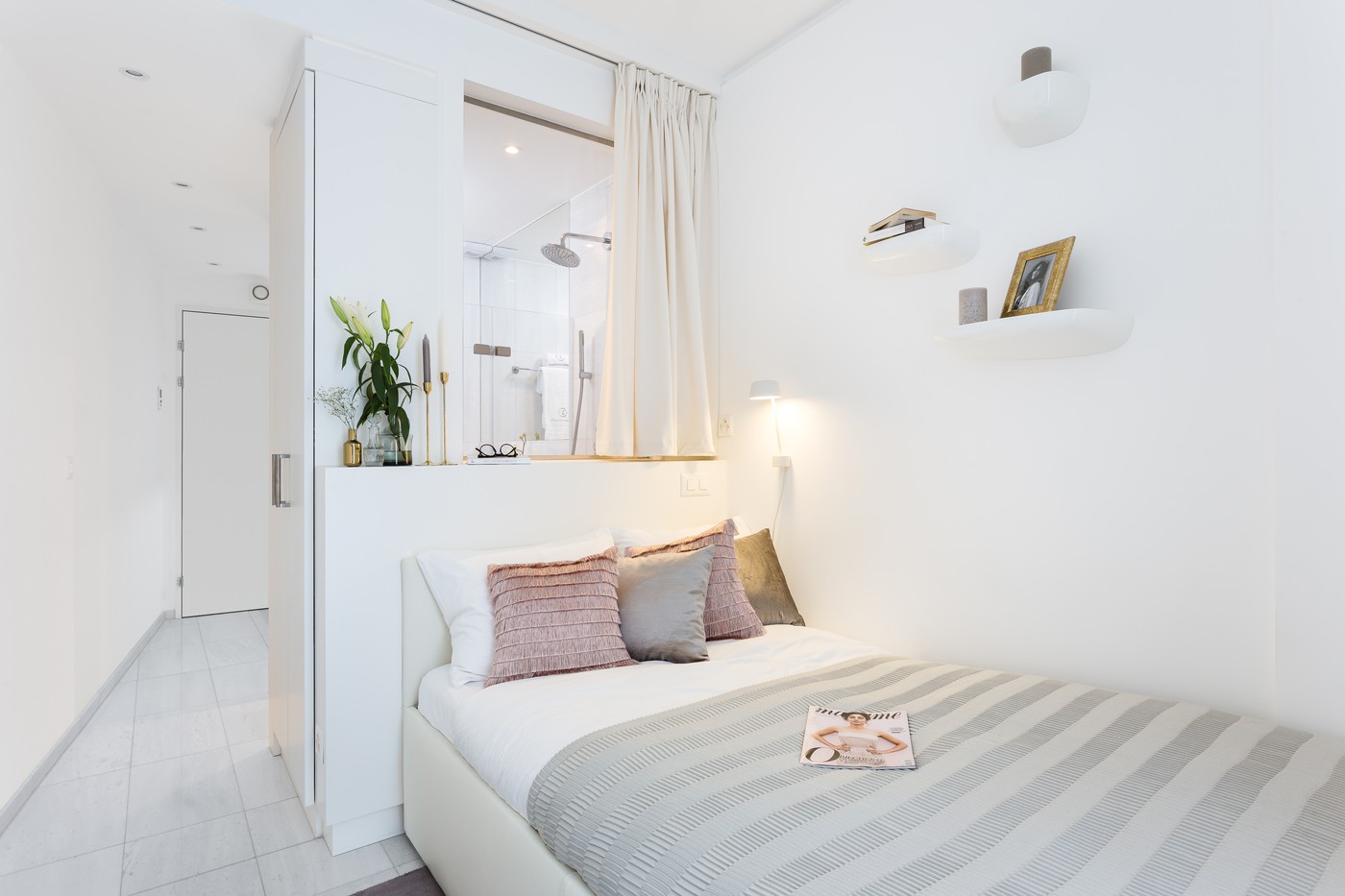 Modern small bedroom with tiled floor, a double bed with a gray and white striped bedspread, a nightstand with magazines, built-in wardrobe, a curtain for the shower, shelves with decorative items, a plant, and a decorative lamp.