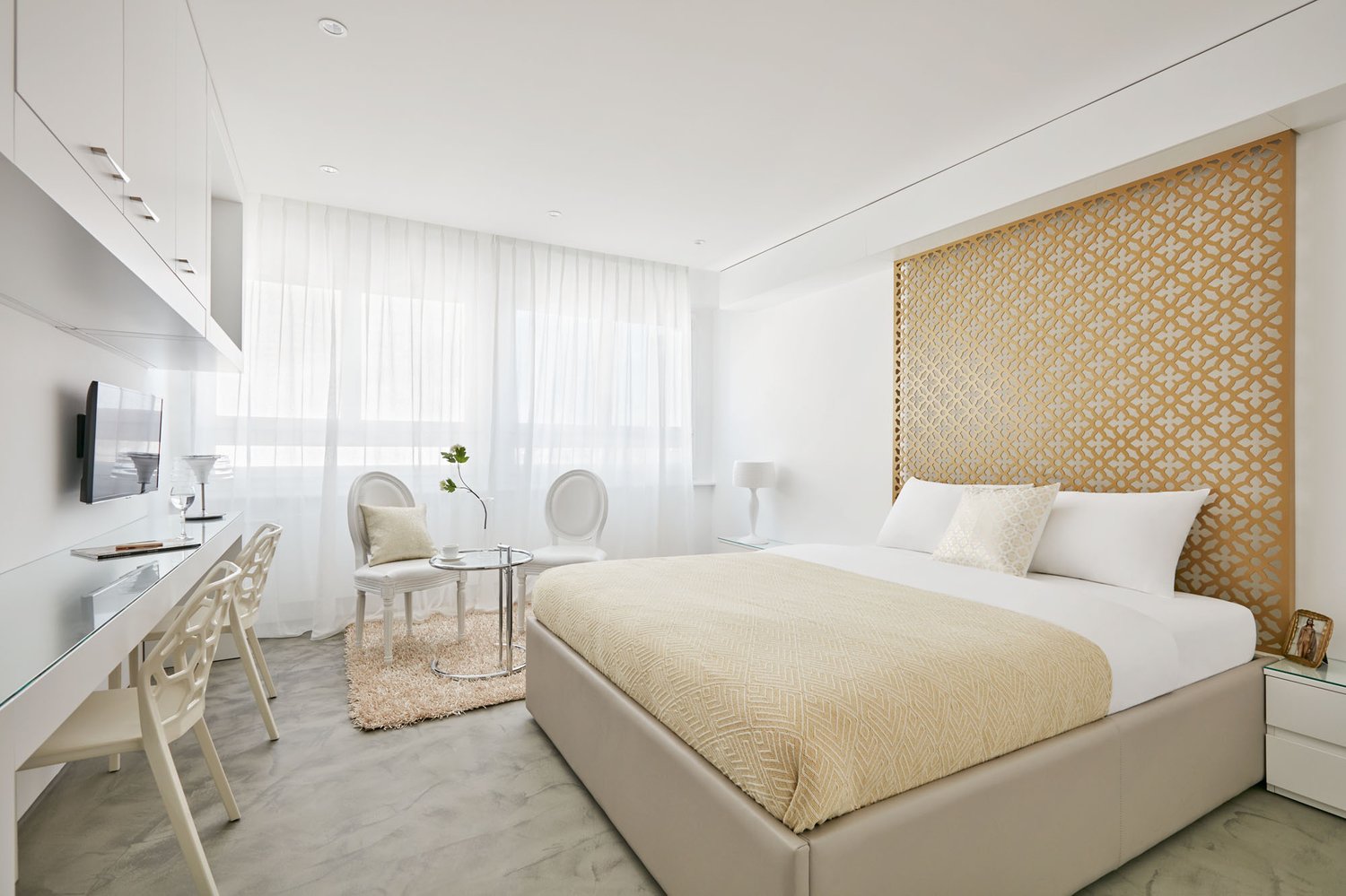 Modern bedroom with beige bed, white headboard, two white armchairs, round glass table, decorative rug, large windows with white curtains, and a desk with a television on the wall.