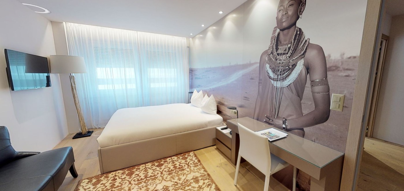 A compact room with a bed, wall mounted television, desk with chair, lamp, sofa, and wall mural.