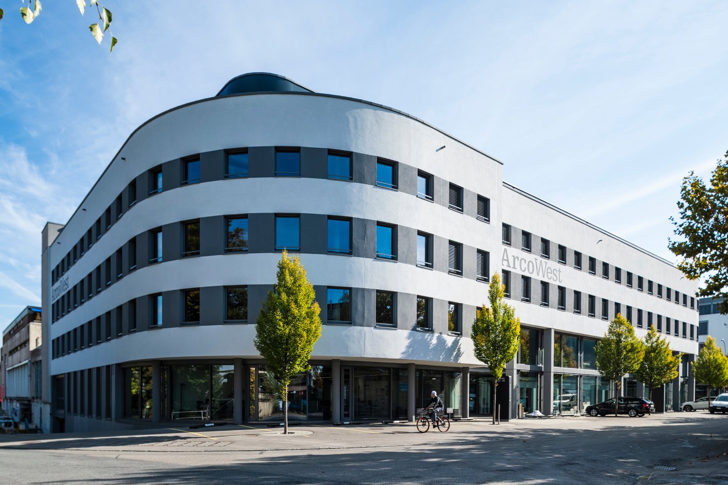 Modern, multi-story office building with a curved facade and glass windows, ArcoWest name sign, trees and cars in front.