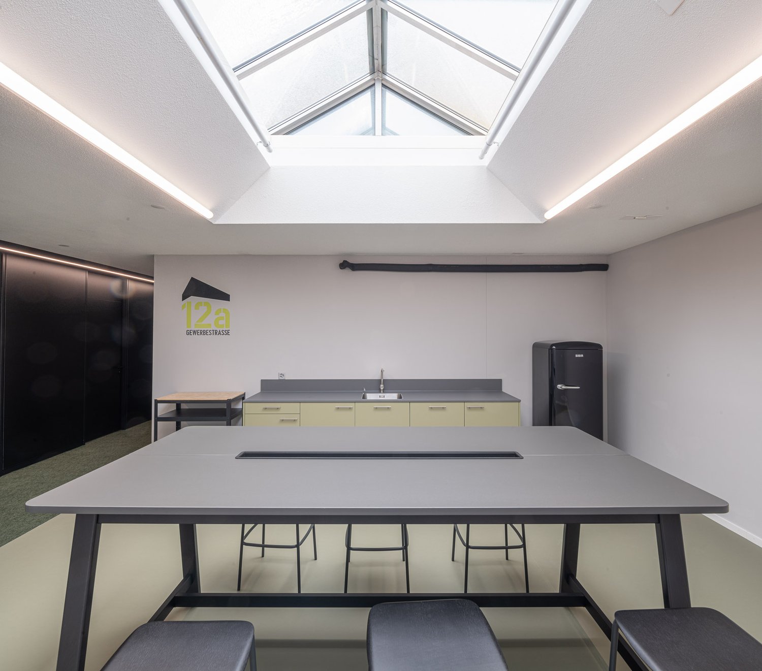Empty kitchen area with a long table, stools, countertop, sink, and a refrigerator.