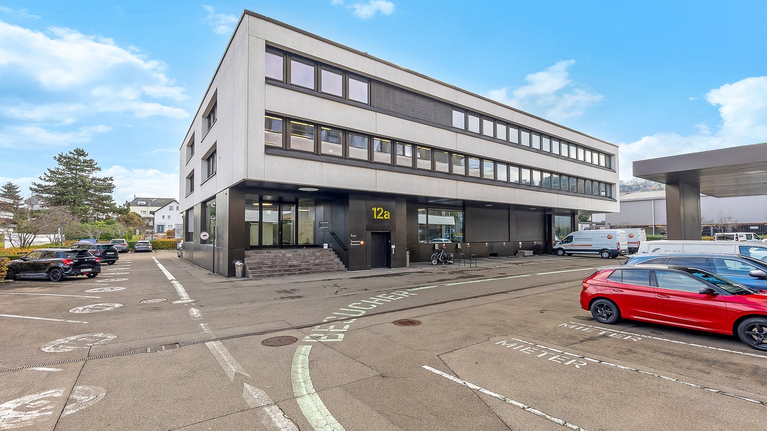 Modern office building, white exterior, multiple windows, entrance with stairs, parking lot, several cars parked