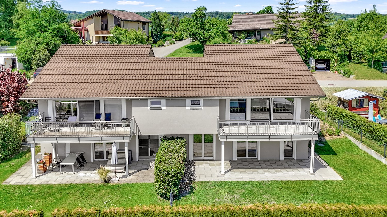 2 story house, brown roof, large windows, two balconies, yard with grass and plants, patio with table, small shed