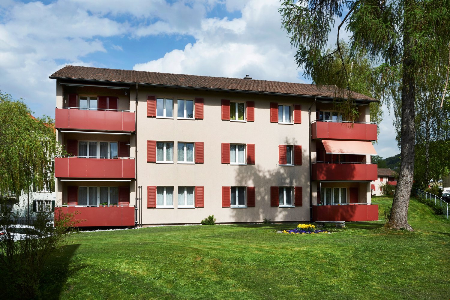 multi-storey building with red balconies and windows, green lawn, tree, and parked cars in the front
