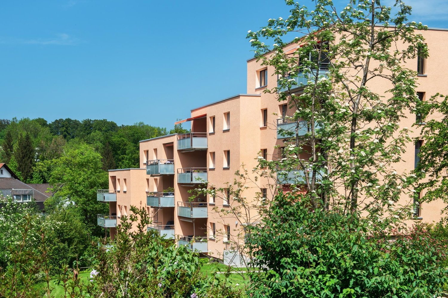 Modern apartment building, beige exterior, multiple balconies, surrounded by greenery, 4 stories