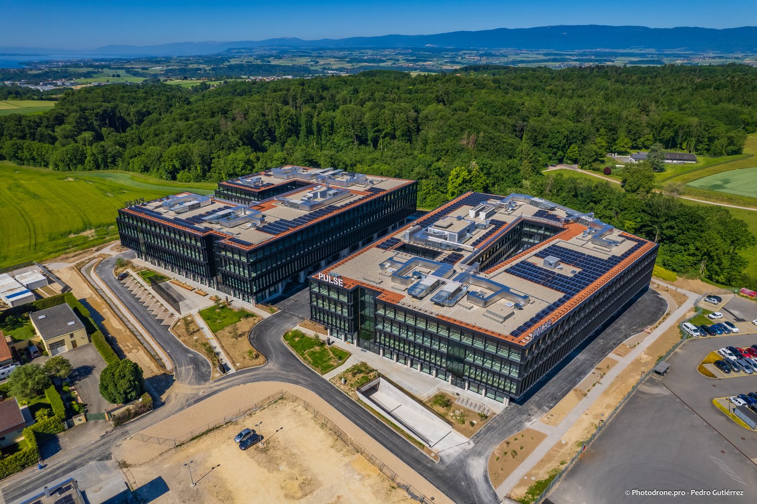 two office buildings with rooftop solar panels, asphalt driveway, parking lot, surrounding trees