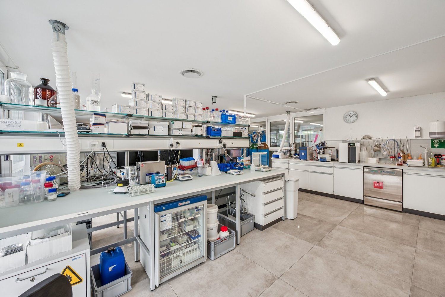 Modern lab interior, stainless steel lab benches, white cabinets, safety signs, various lab equipment and glassware, floor-mounted shelves, stainless steel appliances, and large windows.