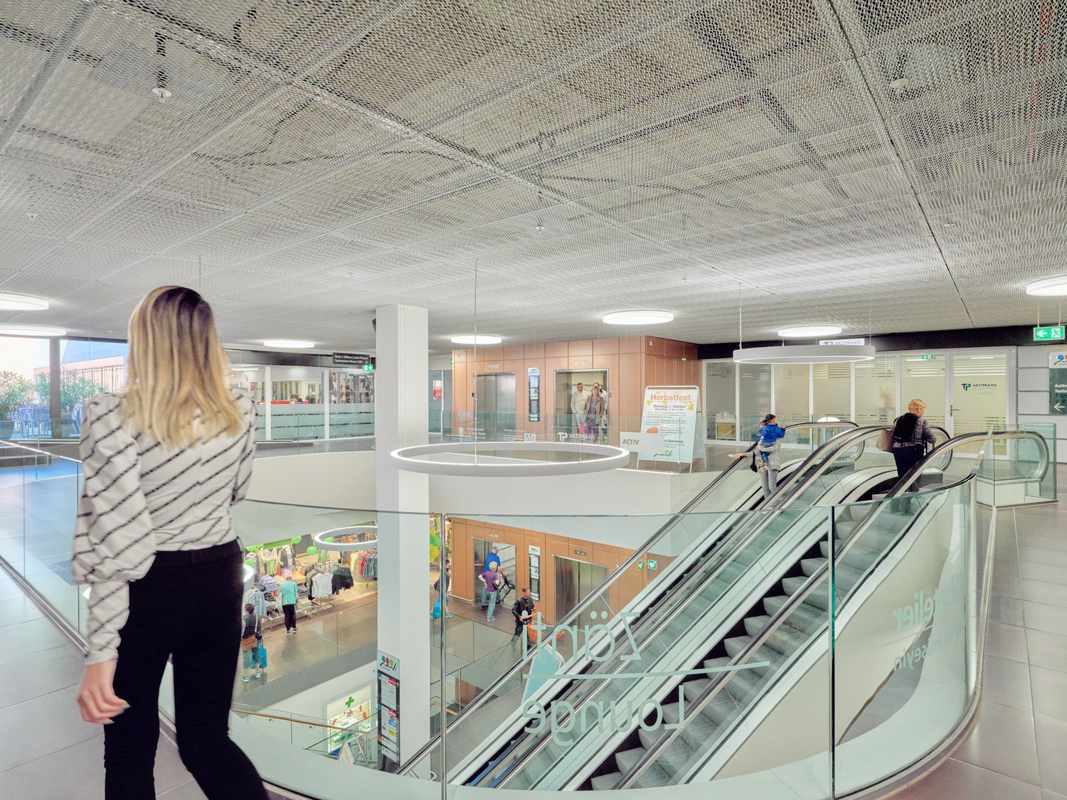 The image shows the interior of a shopping mall. There are escalators, glass walls, and various retail stores visible. The ceiling has a grid-like pattern. A person in a striped sweater is visible in the foreground.