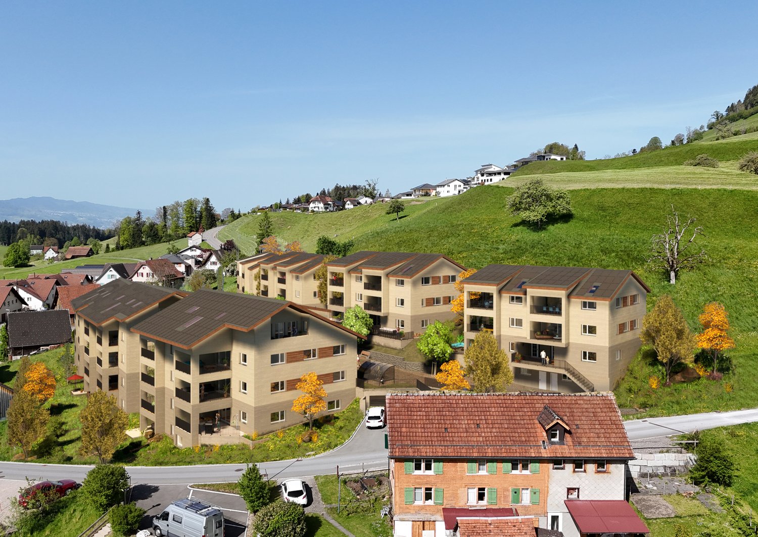 The image shows a residential area with several multi-story apartment buildings nestled in a hilly, forested landscape. The buildings have red tile roofs and appear to be surrounded by parking spaces. There are also some smaller, individual houses visible