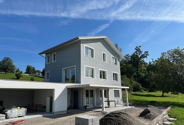 2 story house, grey walls, glass windows, glass doors, entrance with roof, cars park, garage in front, table on the terrace, trees, green field