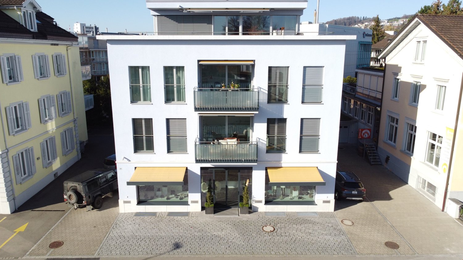 4-story modern white building with large windows, balconies, and retail spaces on the ground floor