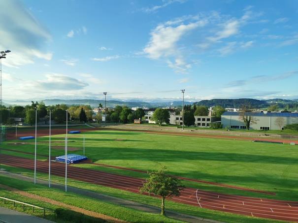 Open green field with track and field poles in the middle, trees on the sides, several buildings in the distance