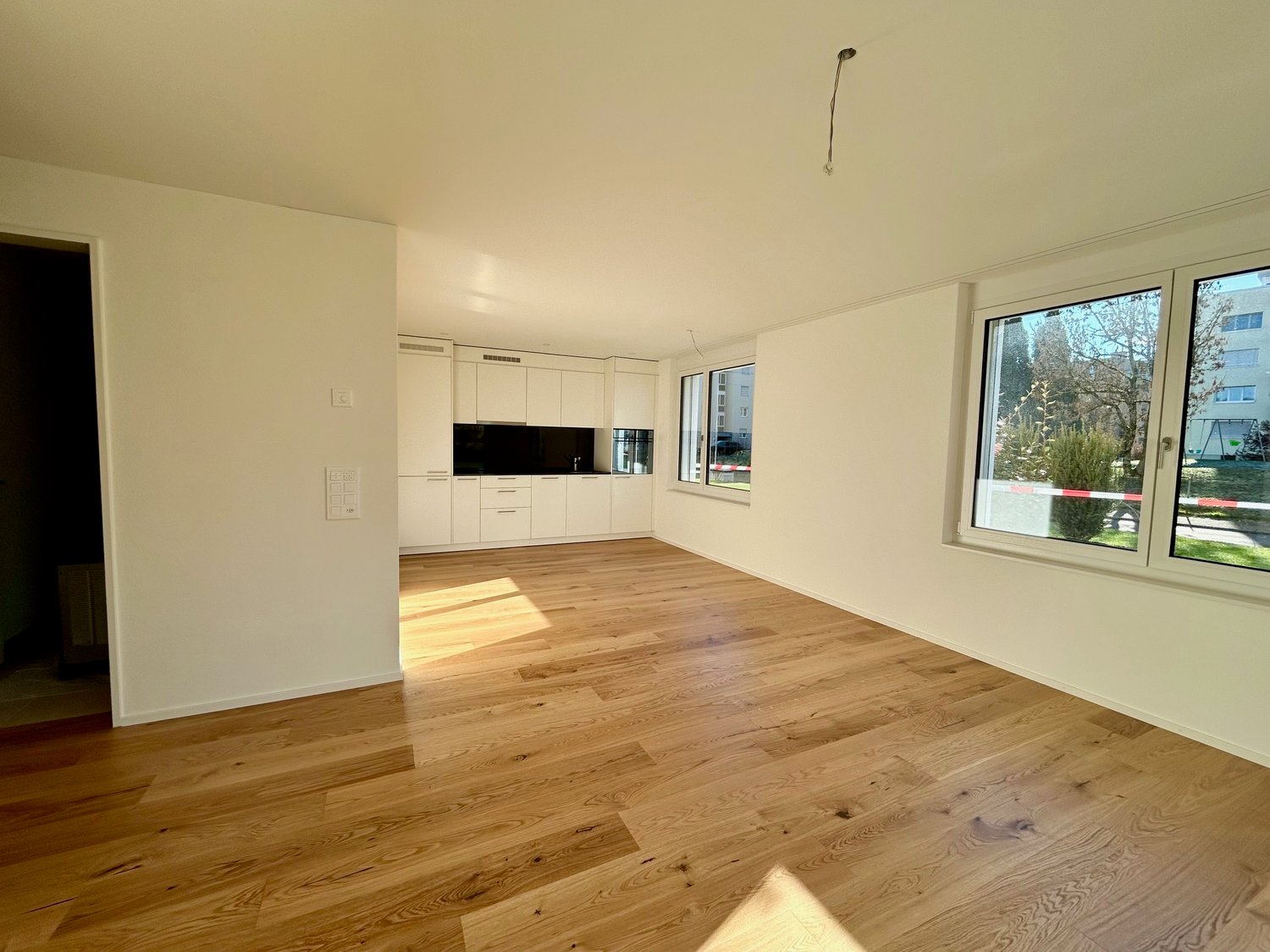 large empty room with white walls, wooden floor, large windows, kitchen area, balcony