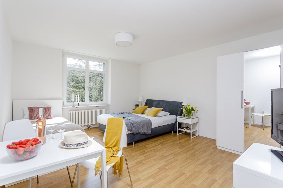 Bright and spacious studio apartment with large windows, hardwood floors, a double bed, a dining table, and a TV stand. The room has a cozy and modern feel with yellow accents and decorative elements.