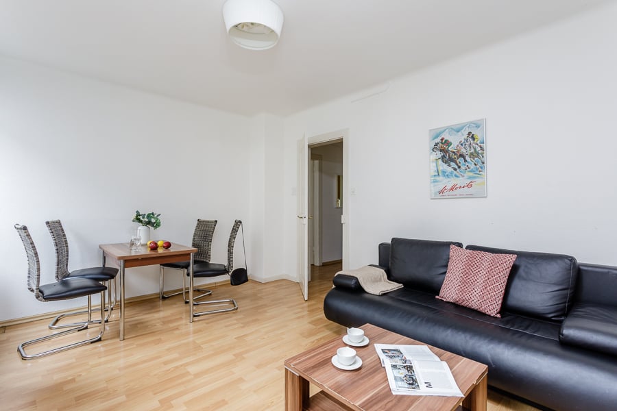 Spacious living room with hardwood floors, white walls, large windows, black leather sofa, wooden dining table with chairs, and a framed poster on the wall.