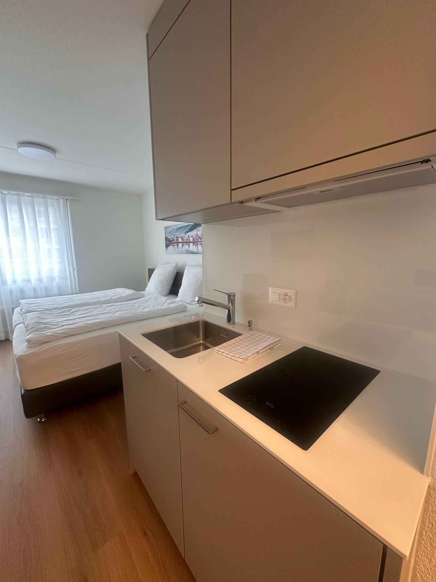 The image shows a studio apartment with a kitchenette area. The kitchenette has a sink, a cooktop, and a dishwasher. There is a bed visible in the background, indicating this is a studio apartment with a combined living and sleeping space.