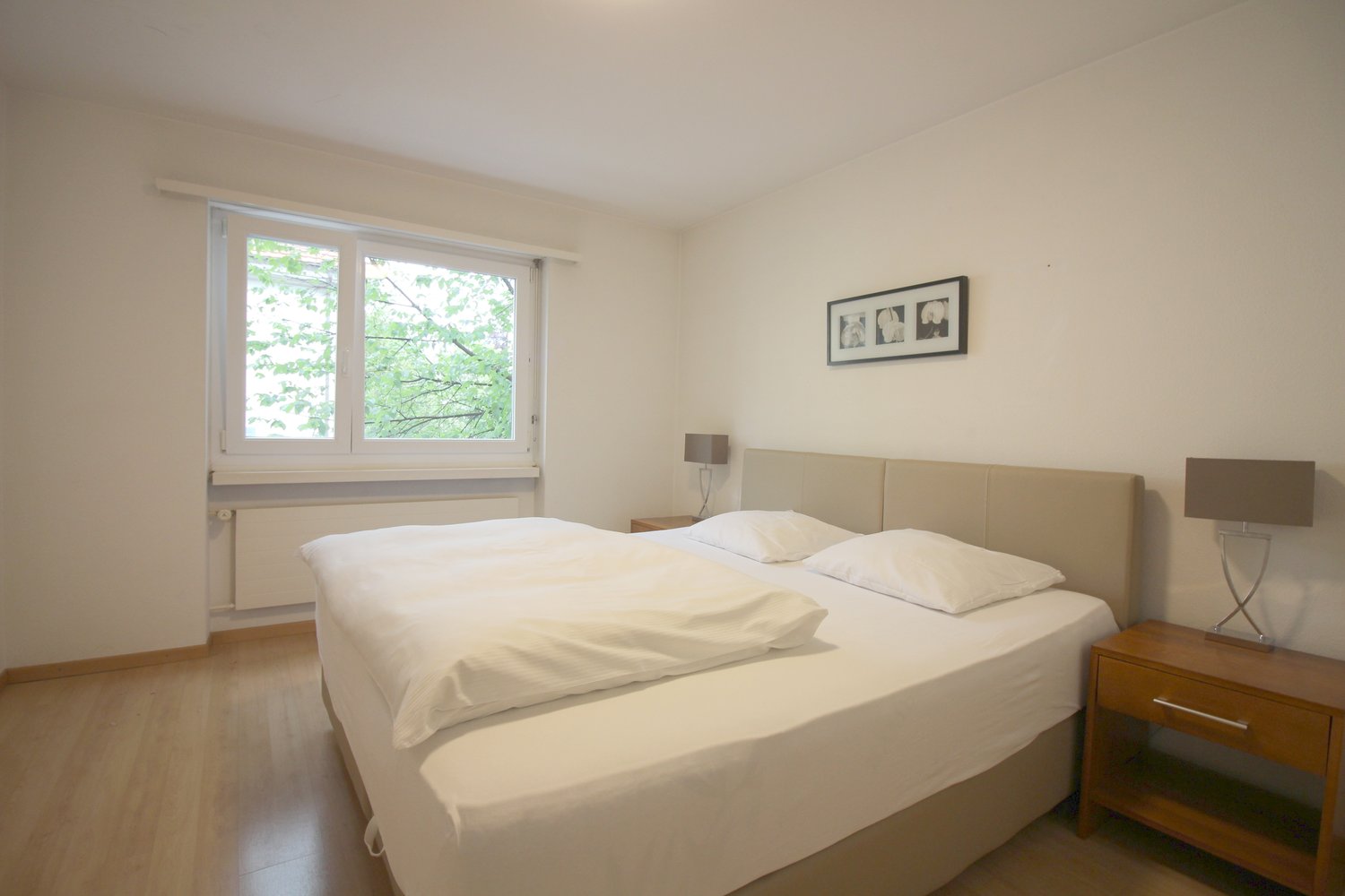 Spacious bedroom with large windows overlooking greenery, white walls, hardwood floors, a double bed with white bedding, a wooden nightstand, and a framed artwork on the wall.