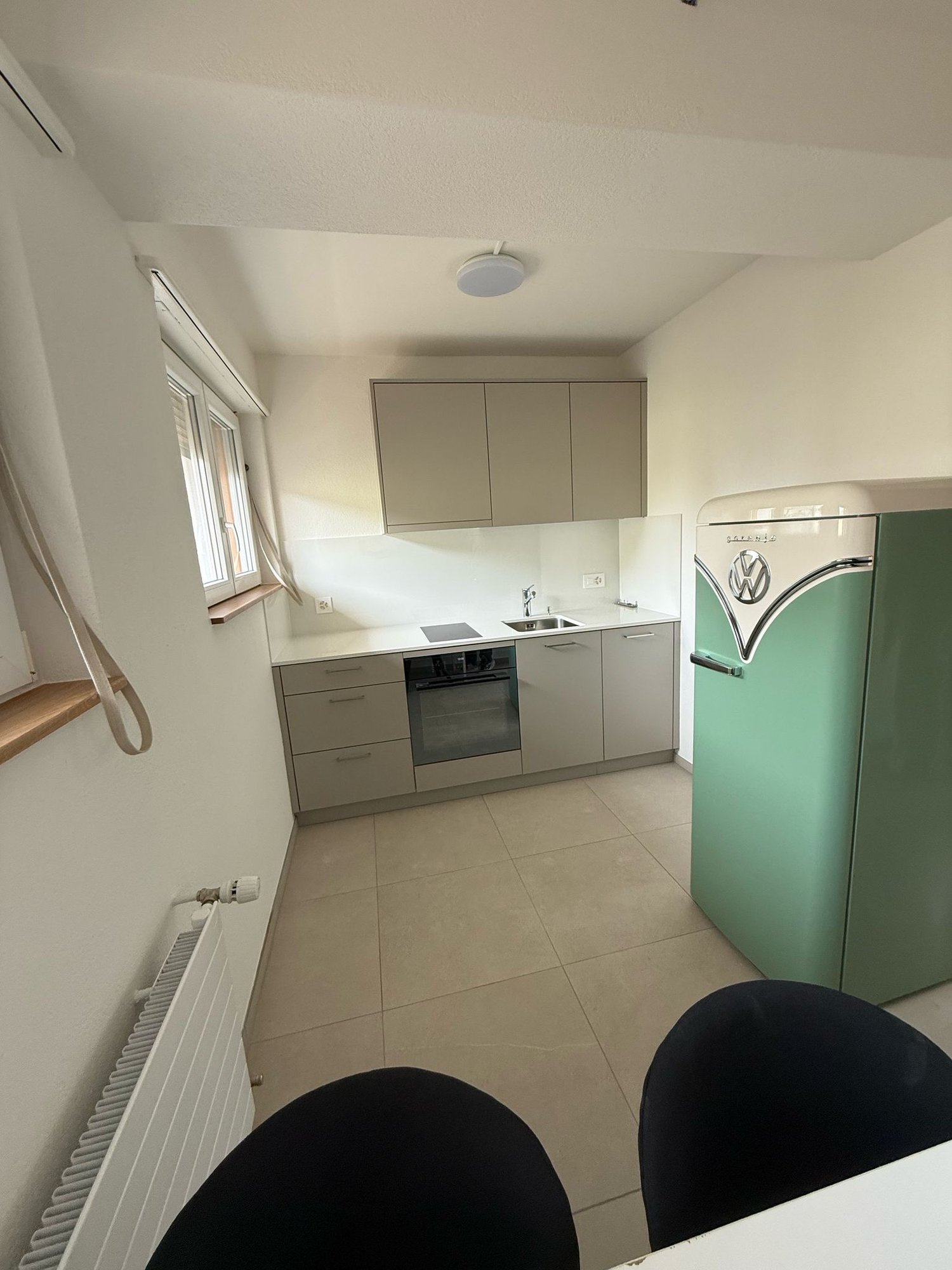 Modern kitchen with gray cabinets, built-in oven, dishwasher, and a retro-style refrigerator in mint green color. The kitchen has tiled flooring and a window.