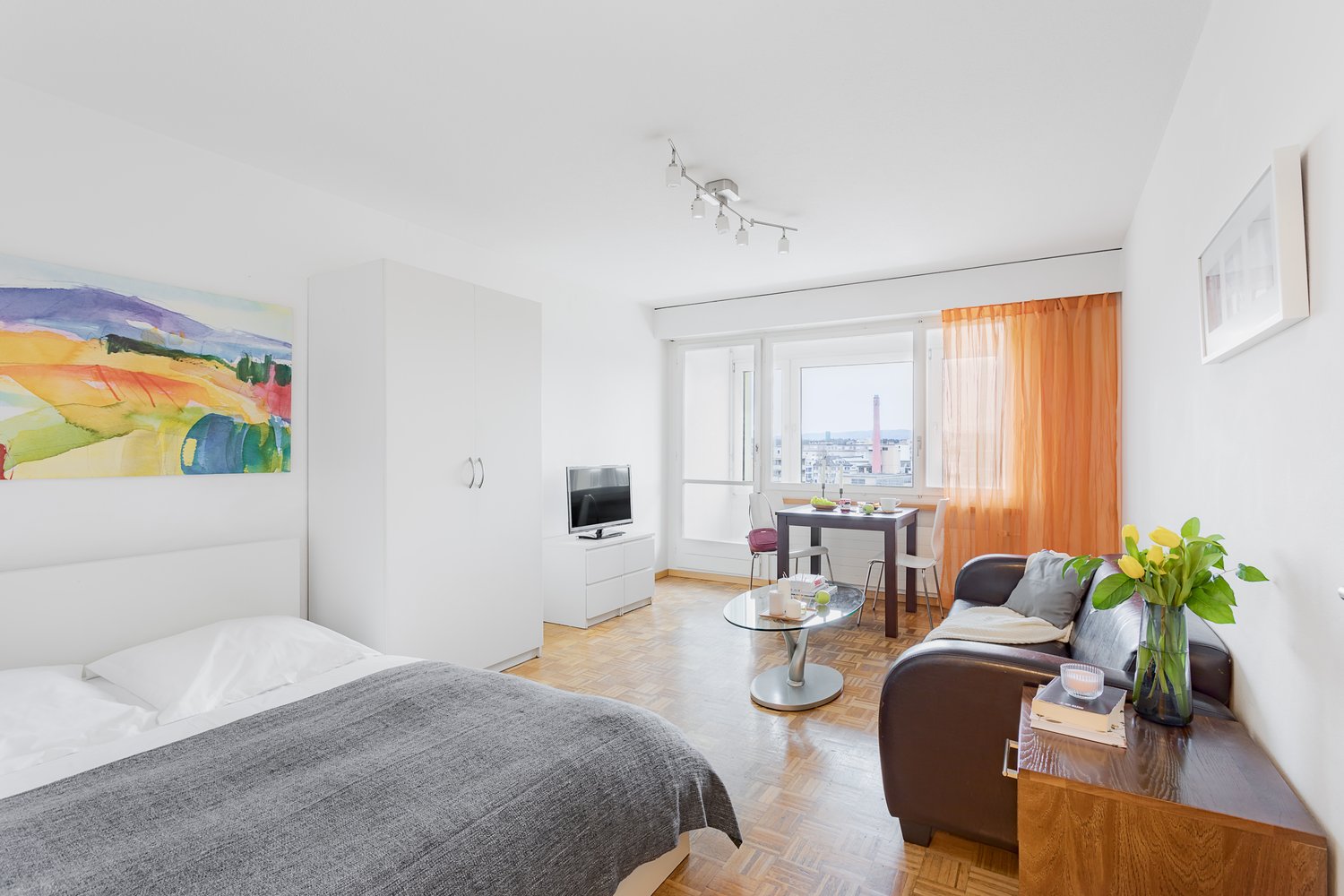 Bright and spacious studio apartment with large windows offering a view of the city. The room features a bed, a sofa, a dining table, and a TV stand. The walls are decorated with a colorful landscape painting. The apartment has hardwood floors and modern