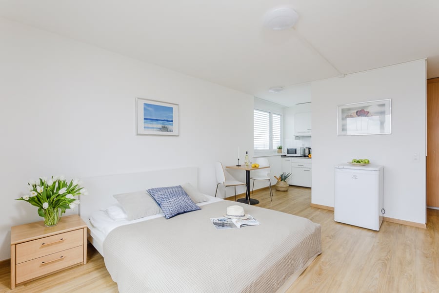 white walls, wooden floor, single bed, pillows, hat, table with flowers, kitchenette, refrigerator, picture frames