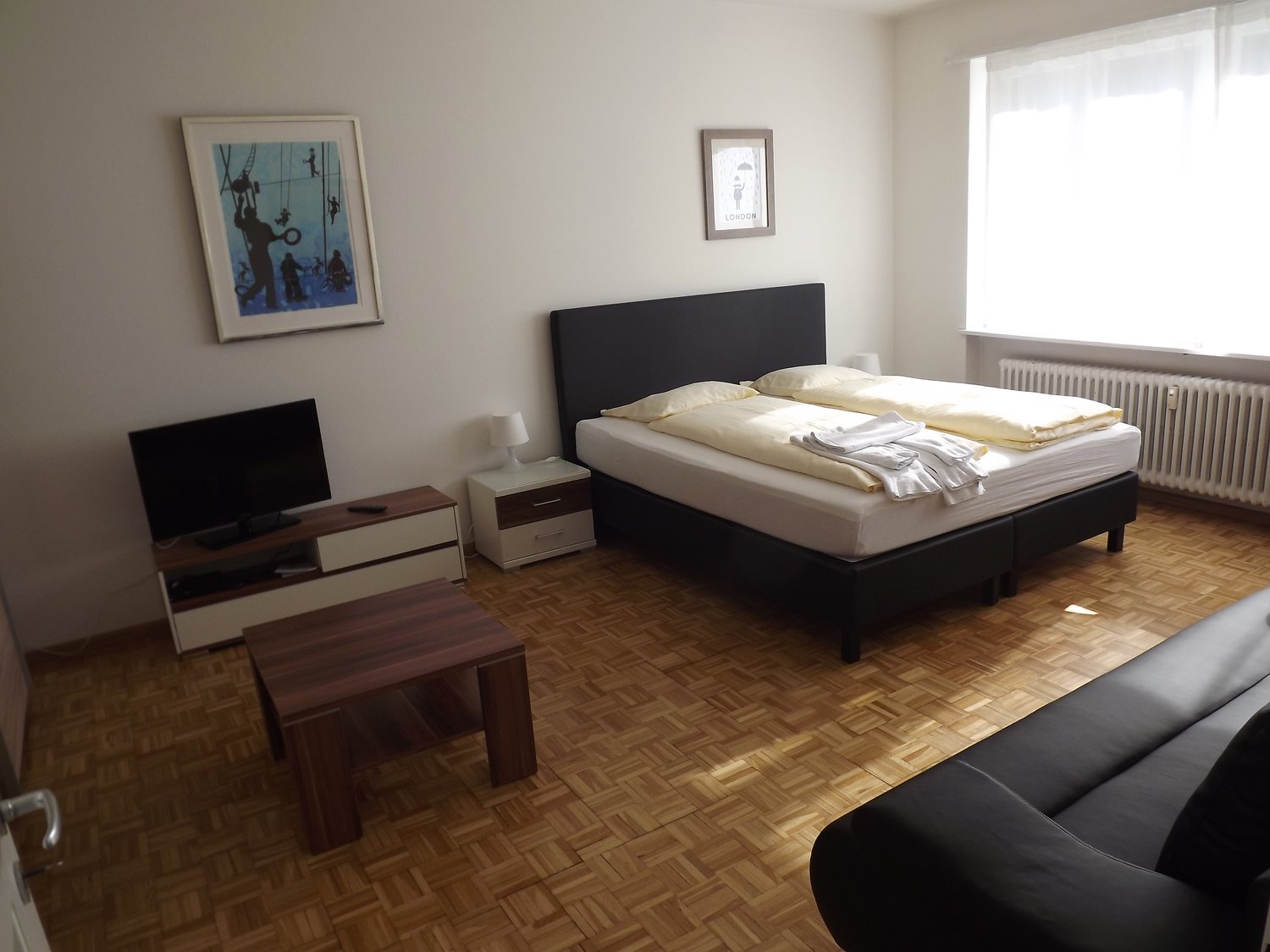 a bedroom with wooden floor, large bed, night table, TV on TV stand, a table, and a couch