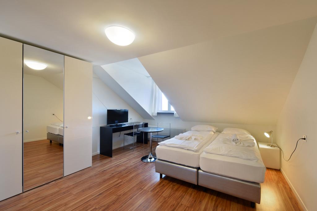 Two double beds, desk with chair, flat screen tv, small table, white walls, wooden floor, lamp, power outlet, mirror on the wall