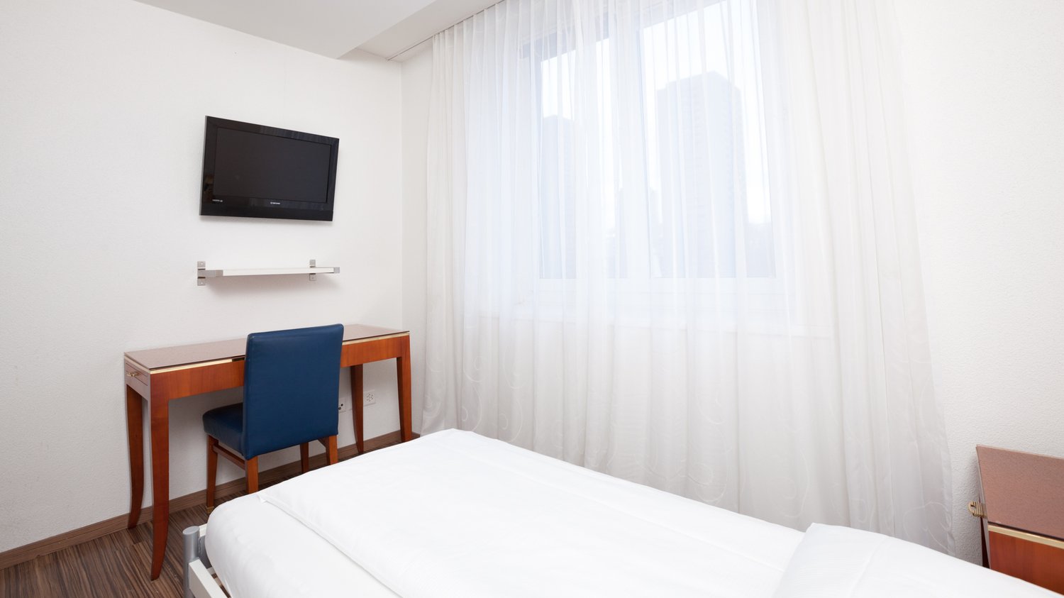 white single bed, wooden desk, blue chair, wall mounted TV, wall outlet, window with curtains