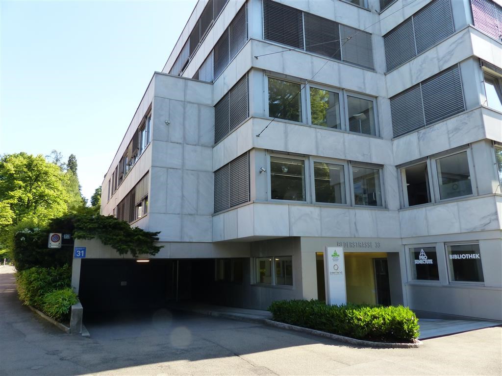 A multi-story office building with a garage entrance, large windows, and a modern concrete and glass facade. The building is surrounded by trees and greenery.
