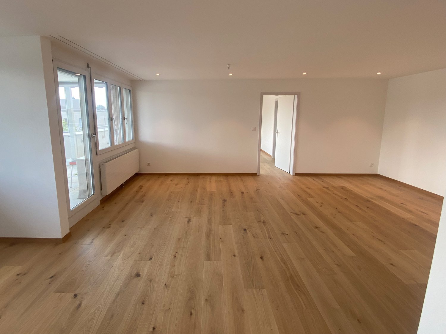 Spacious living room with hardwood flooring, large windows, and a neutral color scheme. The room appears to be empty, ready for furnishing and decor.