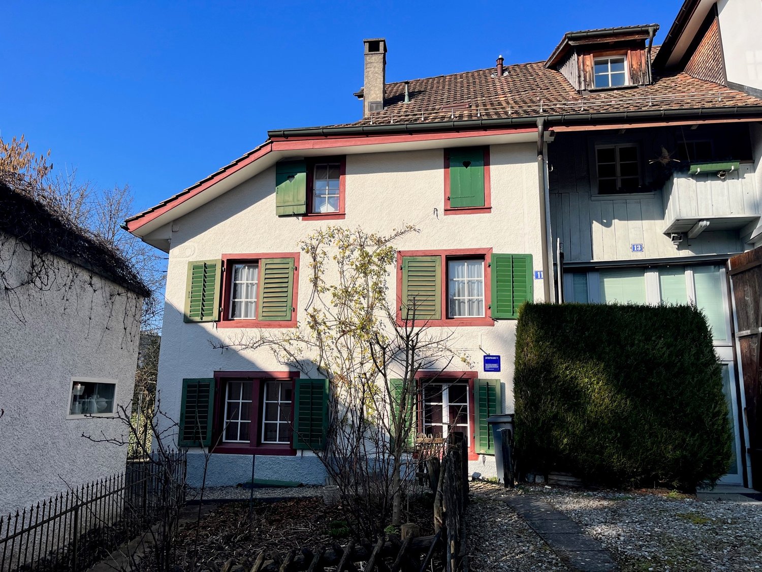 White house with green shutters and red trims, brown roof, several windows, small tree in front, trash bins beside the entrance