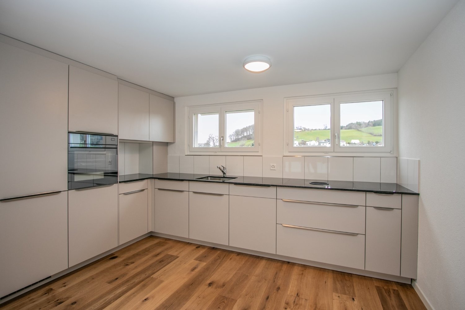 Modern kitchen with white cabinets, black countertops, large windows, under cabinet lighting, built-in oven and sink, wooden floor