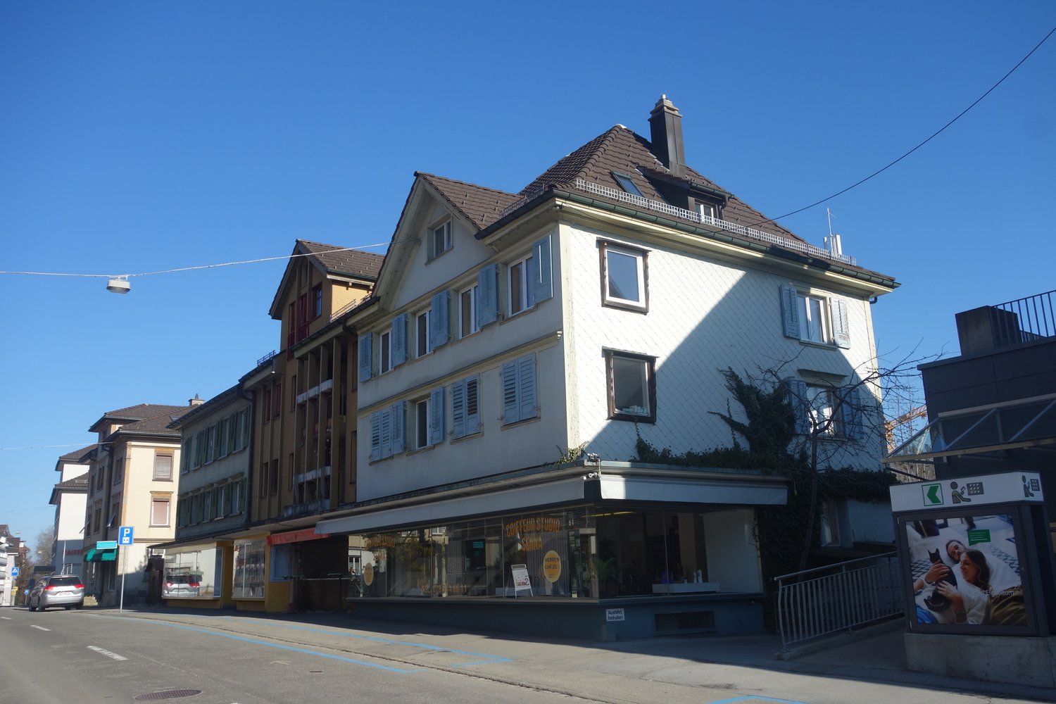Two story building, white exterior, brown roof, several windows, storefronts on the ground floor, metal railings on the sides.