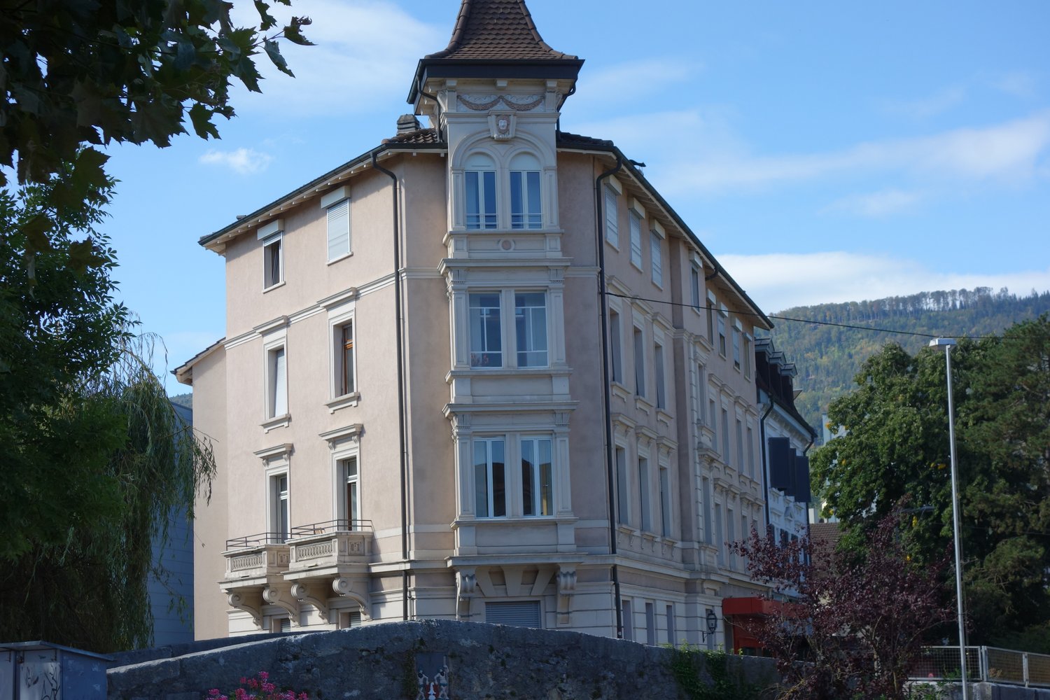 Three-story building, beige exterior, multiple windows, balconies with railings, tower with a pointed roof, surrounded by trees and plants
