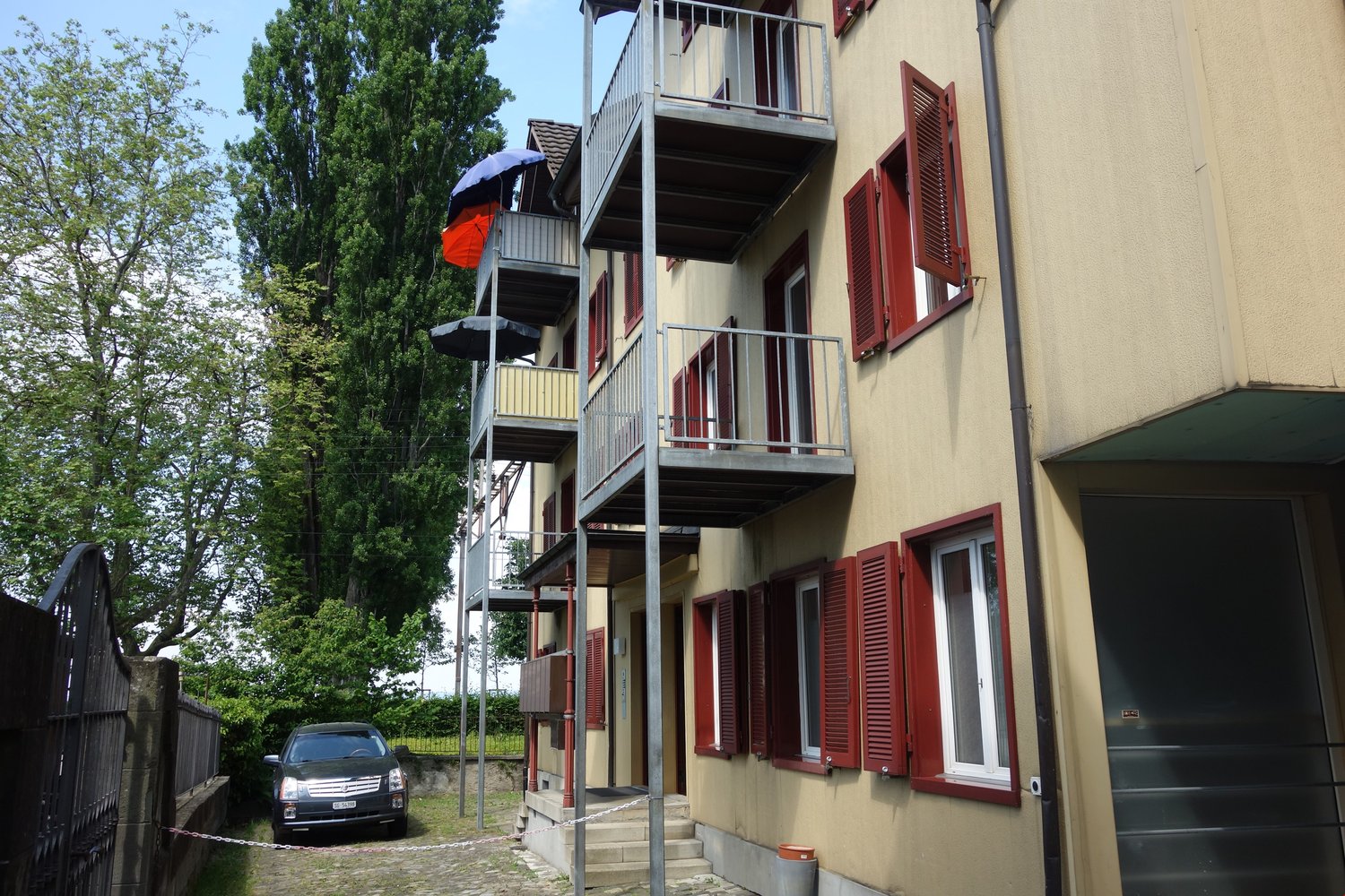 3 story building, yellow wall, red windows, multiple balconies, fence, trees, parking area